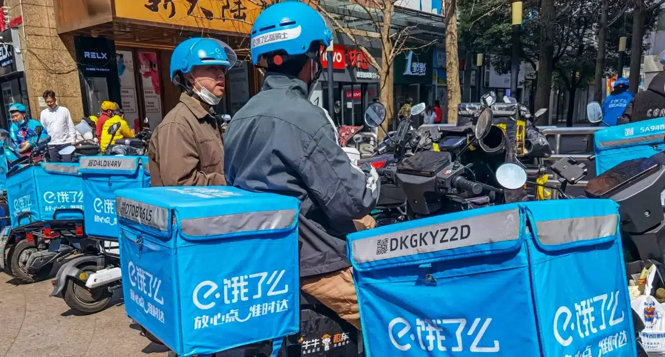 Delivery riders in blue helmets beside scooters loaded with blue insulated delivery boxes on a shopping street